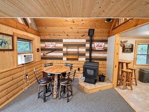 Un interior de cabaña acogedor, con una mesa de madera central, cuatro sillas, un hogar a leña y una ventana con vistas a follaje.