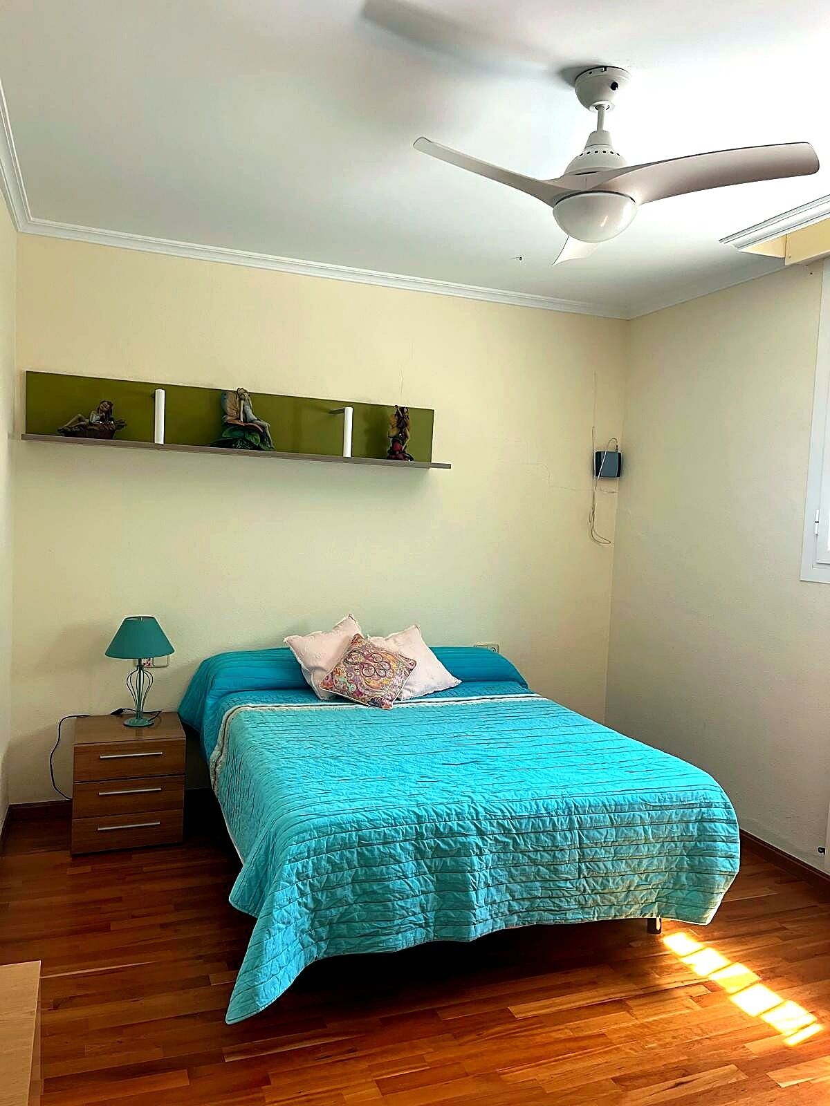 Un dormitorio con cama, mesita de noche, lámpara, ventilador de techo y estanterías en la pared.
