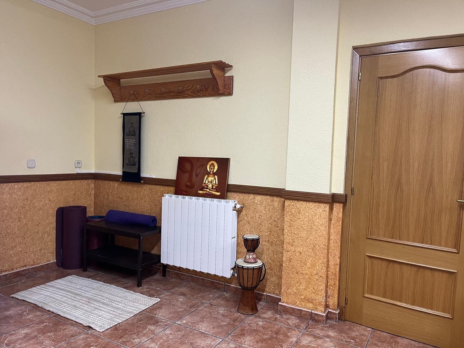 Una habitación con puerta de madera, un radiador blanco, un banco de meditación, una esterilla y un tambor pequeño.