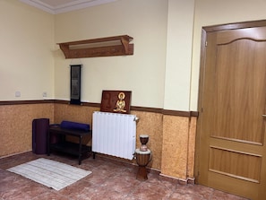 Quarto com porta de madeira, radiador branco, banco de meditação, tapete e um pequeno tambor.