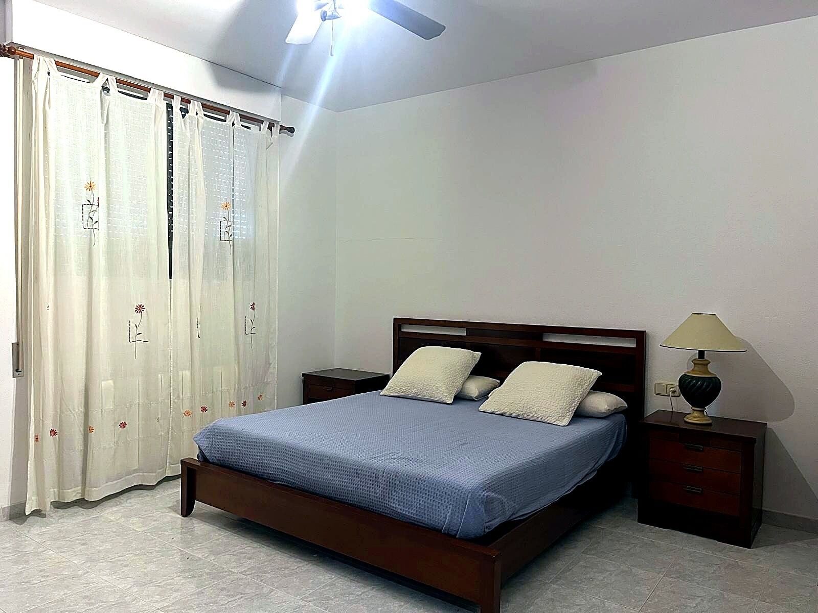 Un dormitorio con cama, cortinas, mesita de noche, lámpara y ventilador de techo.