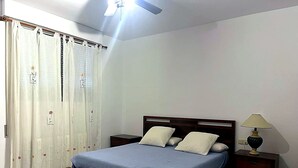 Une chambre à coucher avec un lit, des rideaux, une table de chevet, une lampe et un ventilateur de plafond.