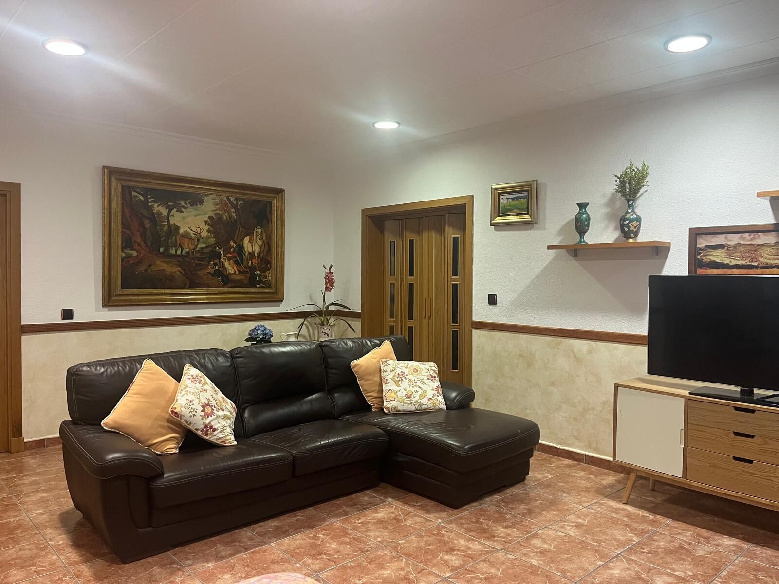 Una sala de estar con un sofá de cuero negro, un televisor sobre un mueble bajo y un cuadro en la pared.
