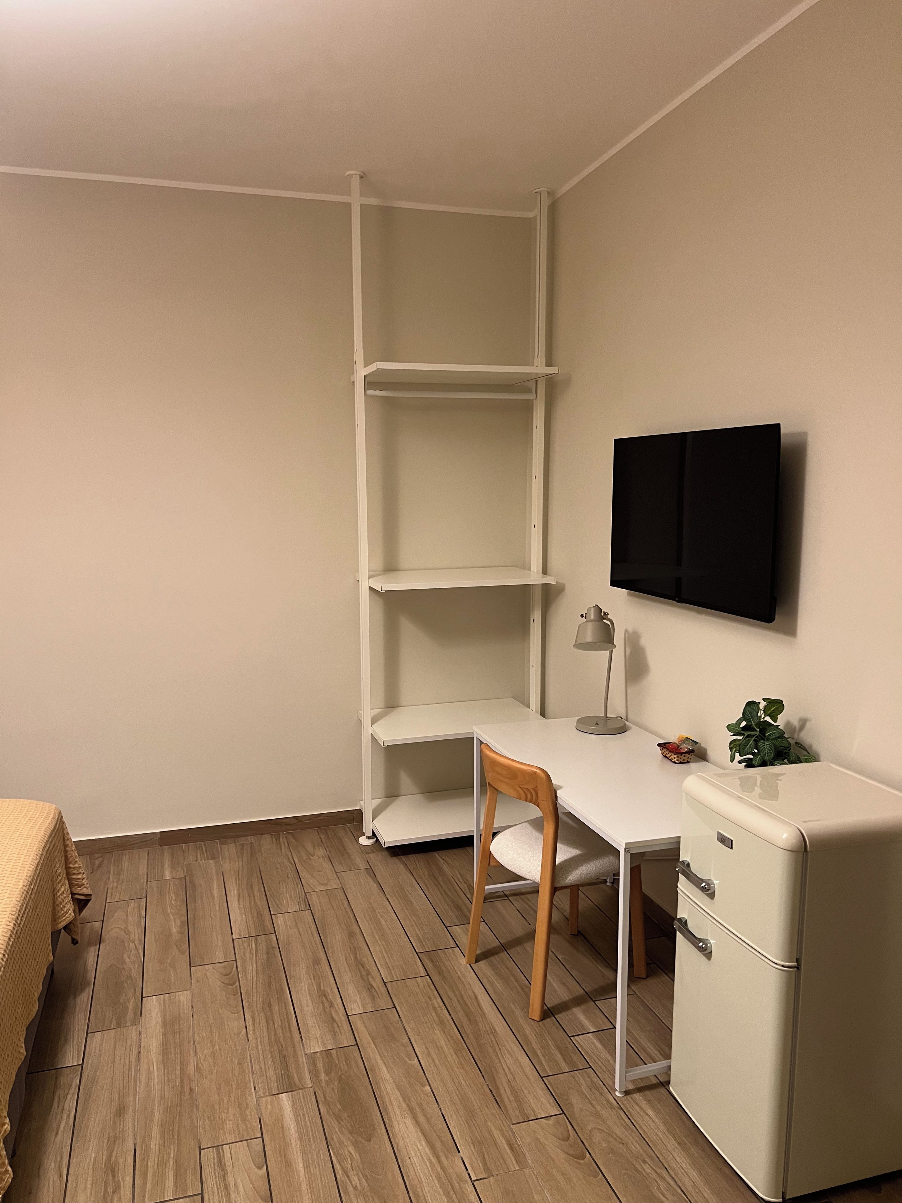 Habitación de esquina con escritorio blanco, una silla, un mueble blanco pequeño y un televisor montado en la pared.