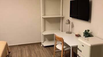 A corner room with a white desk, a chair, a small white cabinet, and a television mounted on the wall.