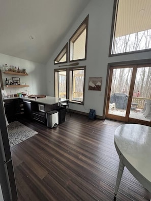 A spacious kitchen with a large island, wooden flooring, and a sliding glass door leading to a backyard. - Cozy modern family build in the woods w/ hot tub! (Cobbs Lake Preserve)