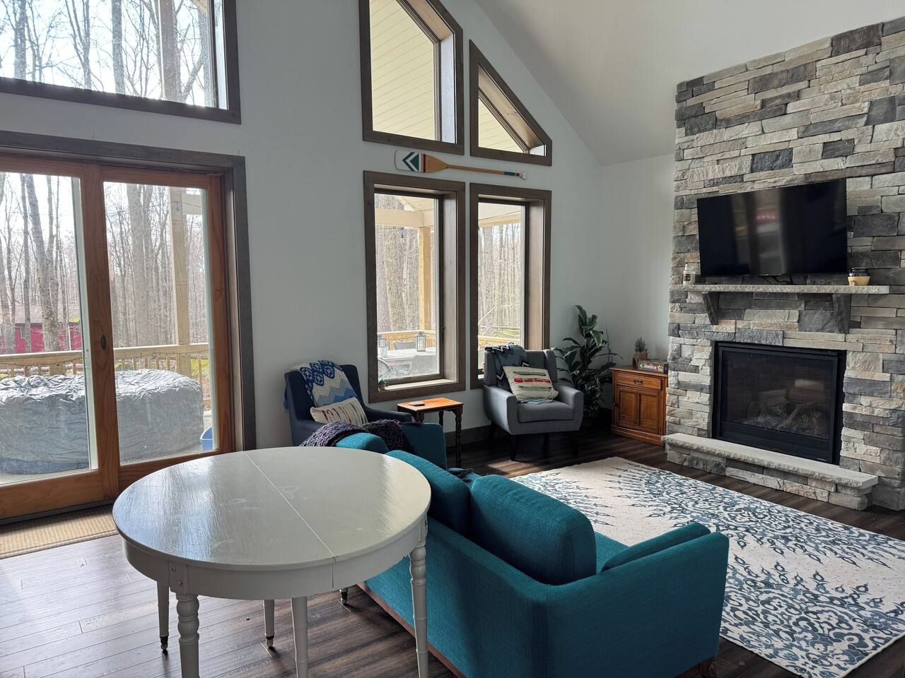 A living room with a stone fireplace, a television mounted above it, and a large window with a view of trees.