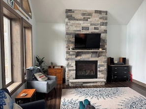 A living room with a stone fireplace, a television, a sofa, a coffee table, and a patterned rug. - Cozy modern family build in the woods w/ hot tub! (Cobbs Lake Preserve)