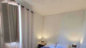A bedroom with a bed, wooden flooring, a headboard with a textured pattern, two bedside lamps, and a window with curtains. - Pousada Casa Hospedaria (Paraty)