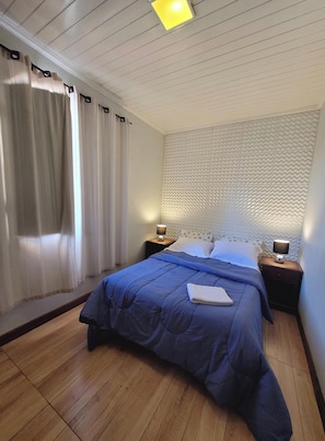 Ein Schlafzimmer mit einem Bett, Holzboden, einem Kopfteil mit strukturierter Oberfläche, zwei Nachttischlampen und einem Fenster mit Vorhängen.
