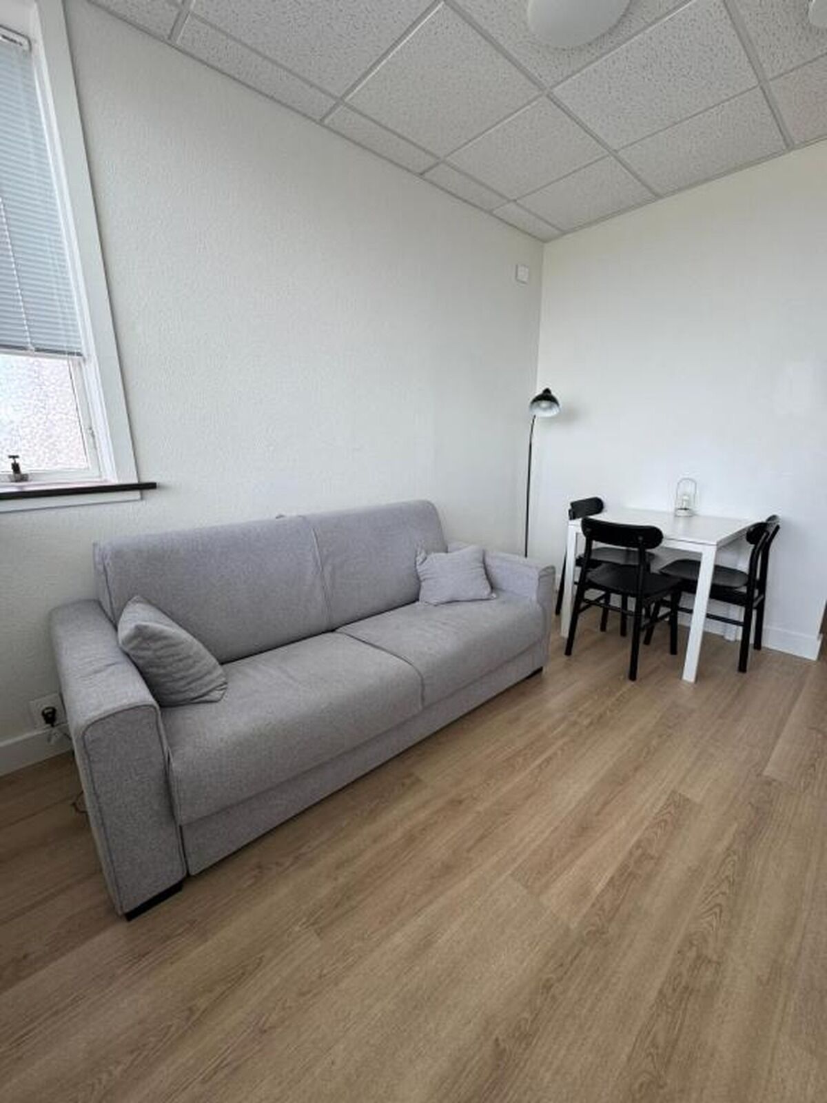 Ein Zimmer mit einer grauen Couch, einem kleinen Tisch mit Stühlen, einer Stehlampe und einem Fenster mit Jalousien.