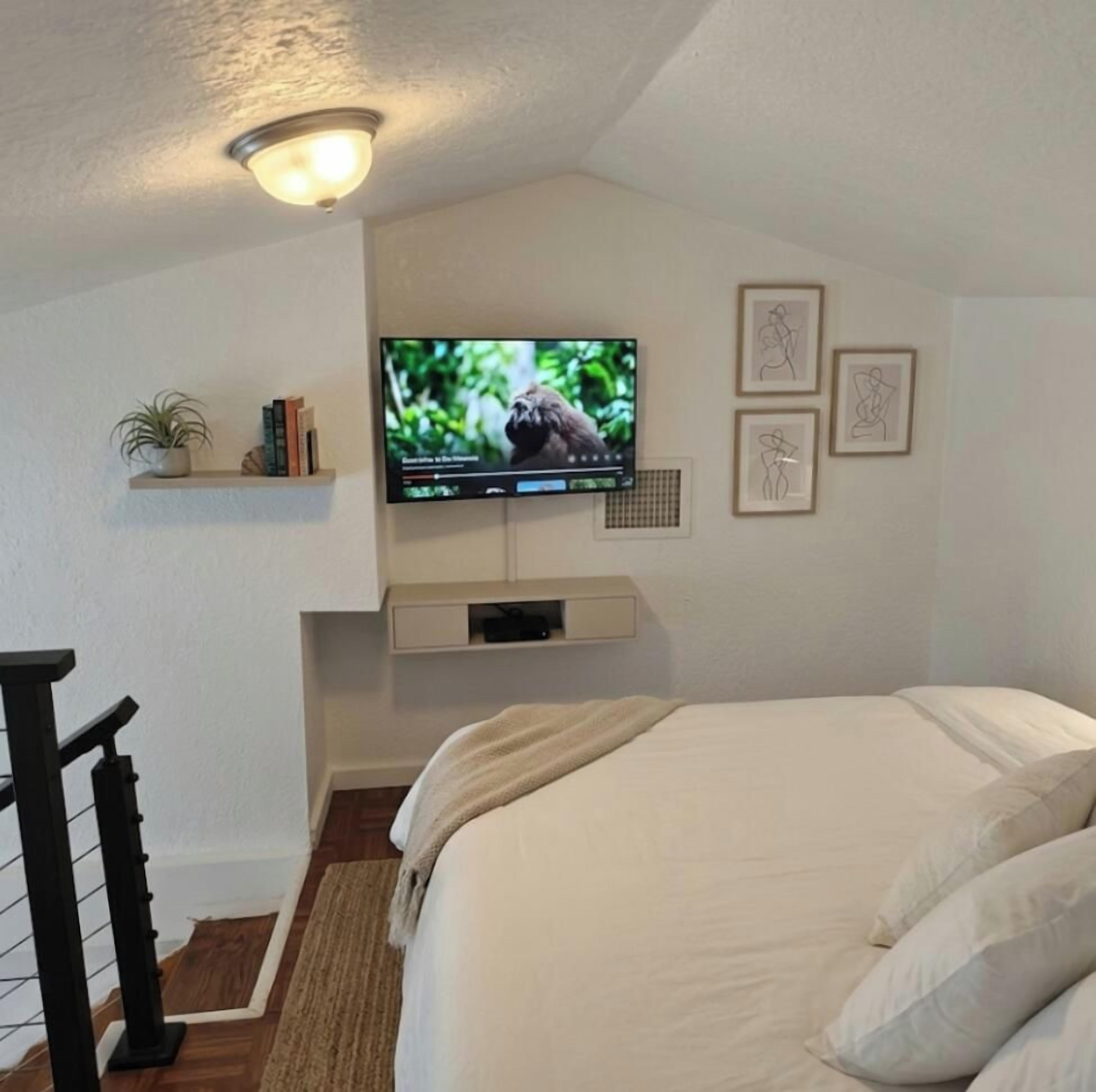 A bedroom with a bed, a television mounted on the wall, a small shelf with books and a plant, and a staircase.