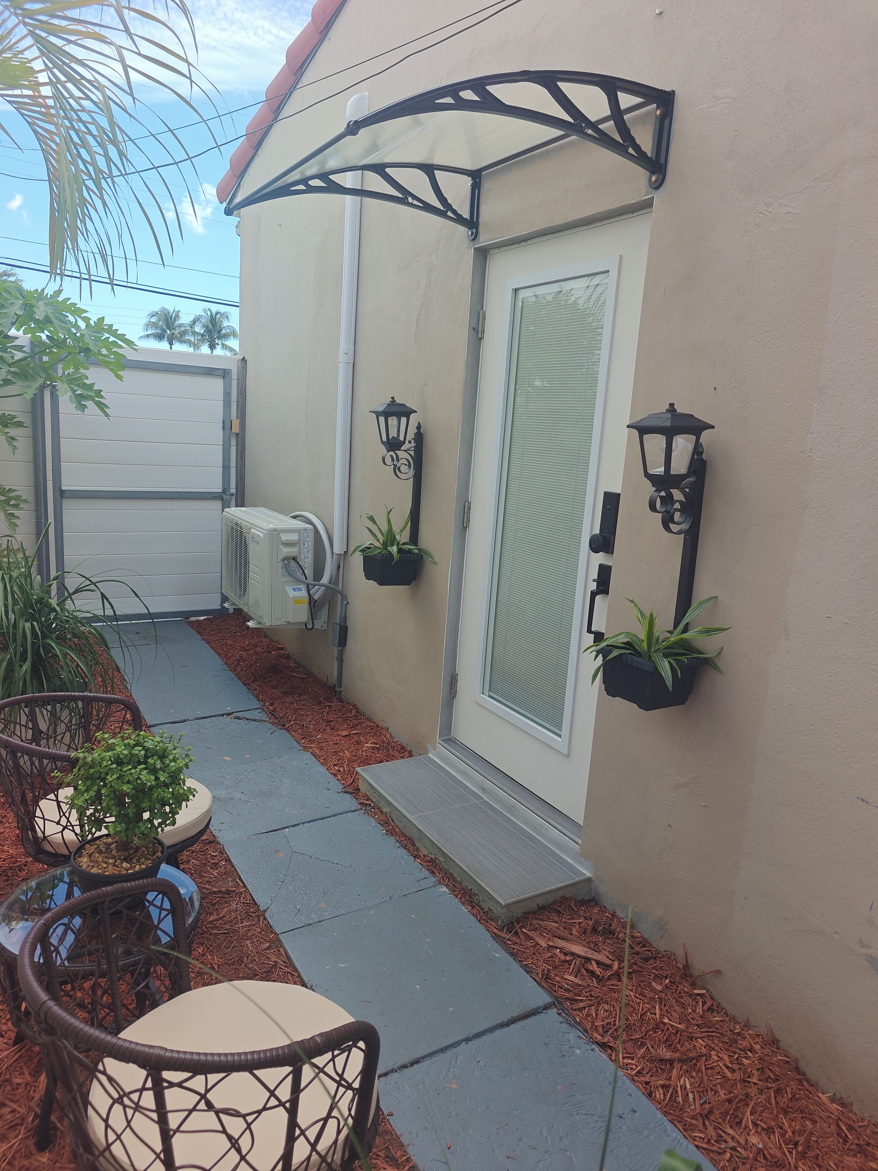 A residential exterior with a covered porch, potted plants, a door with a glass panel, and a pathway leading to a door.