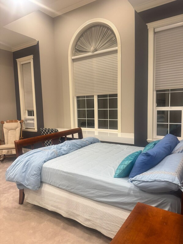 Une chambre avec un grand lit, deux fenêtres équipées de stores, une chaise et un radiateur.