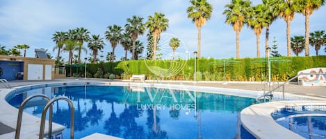 A swimming pool with a clear blue water surface, surrounded by palm trees and a well-maintained hedge.
