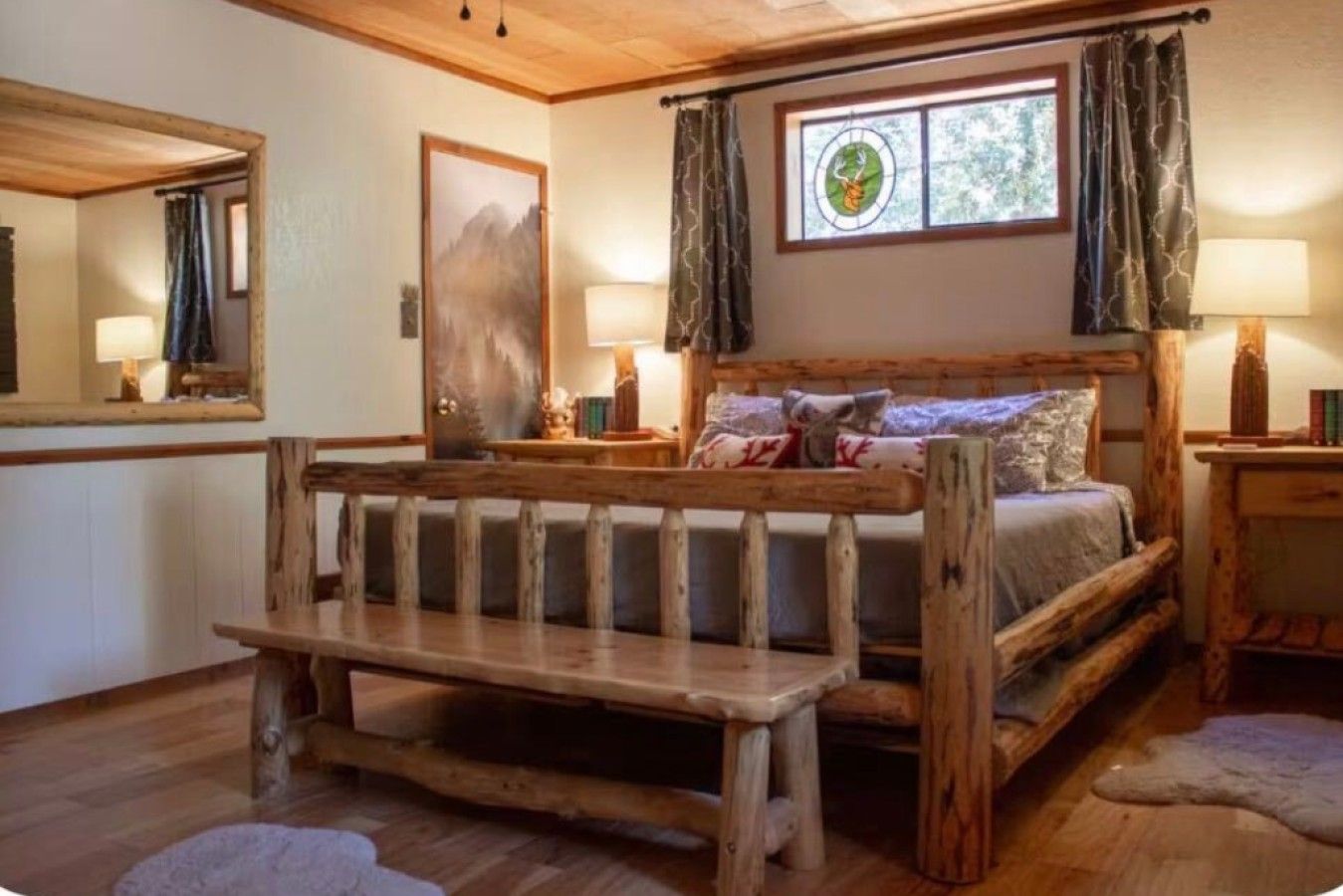 A rustic bedroom with a wooden bed, a bench, a window with curtains, and a mirror.