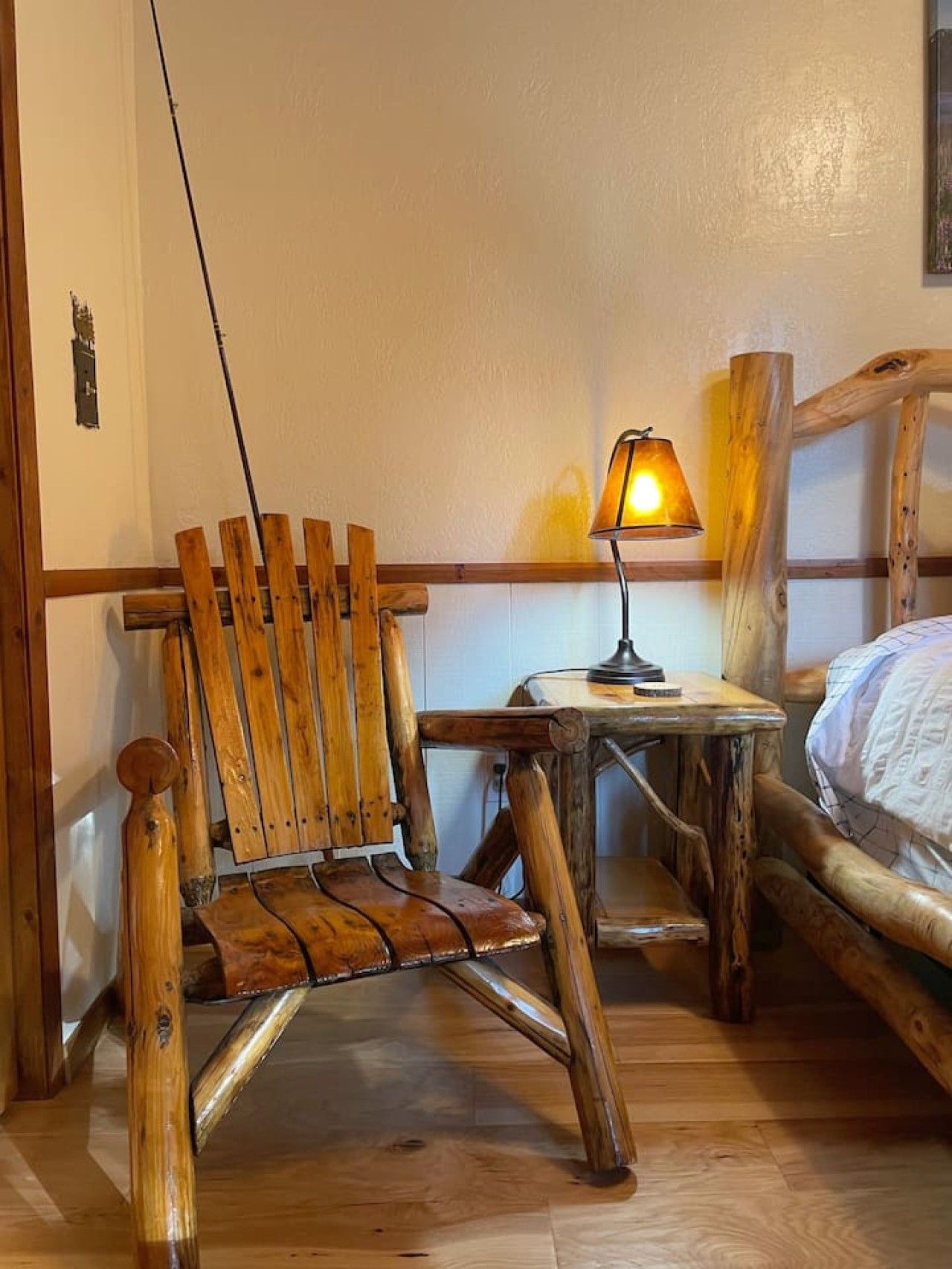 A wooden chair, a small table with a lamp, and a bed in a room with wooden flooring and walls.