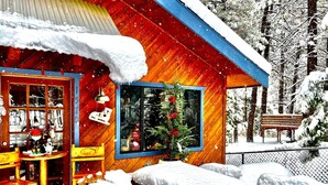 Una cabaña de madera con exterior cubierto de nieve, una ventana con un árbol de Navidad y un banco al fondo.