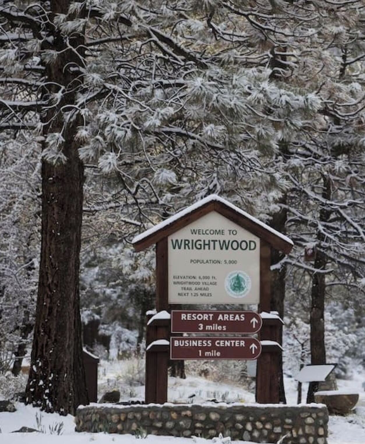 Un panneau indicateur en bois, avec un message d’accueil pour Wrightwood, indiquant une population de 5 000 habitants, une altitude de 1 800 mètres et un sentier qui se trouve plus loin.