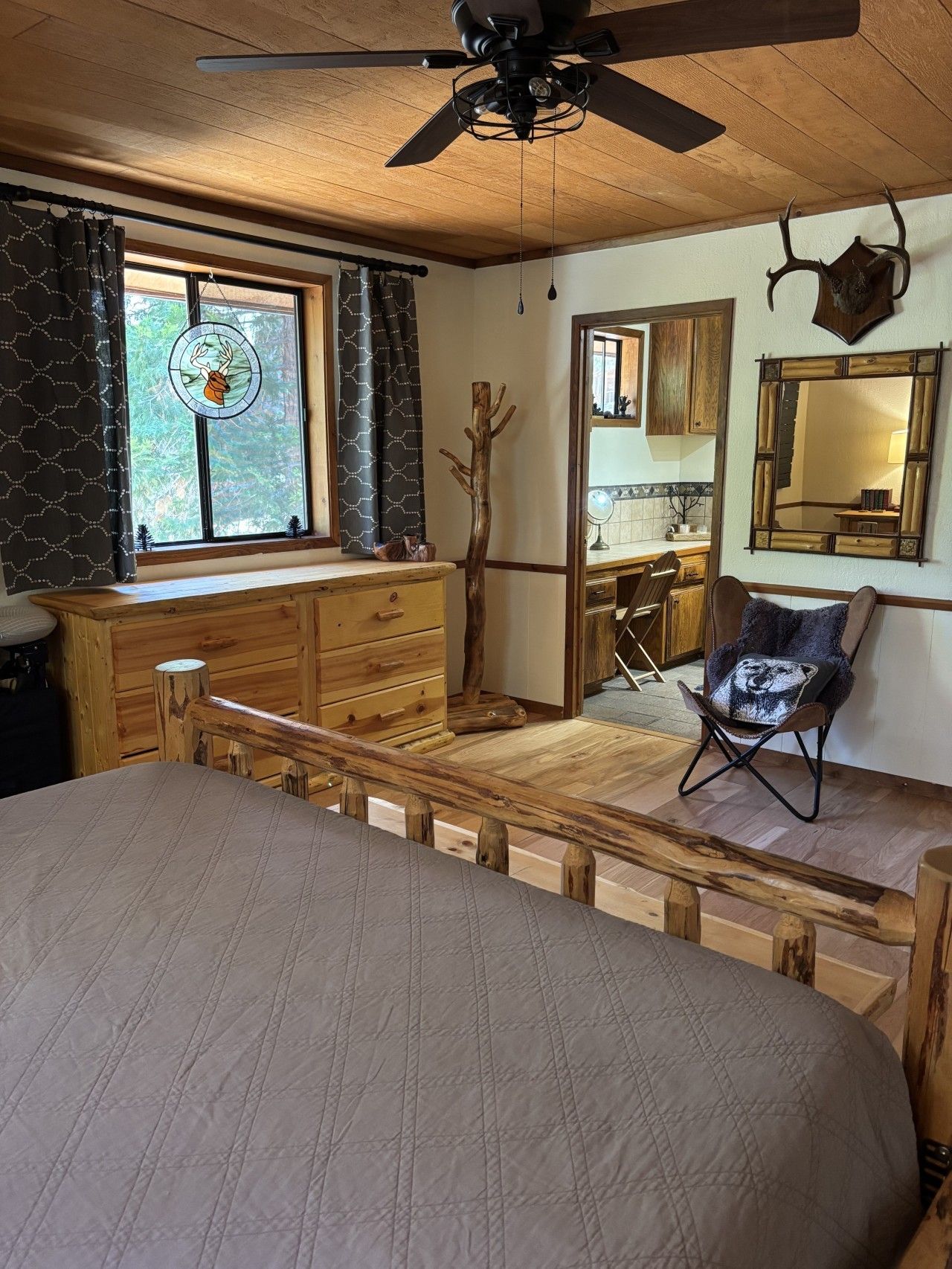 A room with a bed, a wooden bench, a chair with a bear pillow, a ceiling fan, a window with curtains, and a mirror on the wall.