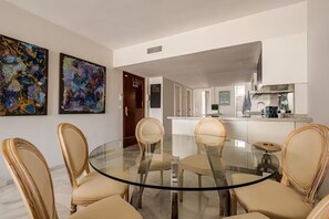 A dining area with a glass table surrounded by chairs, a kitchen in the background, and abstract paintings on the walls. - Cosy Apartment inside Puerto Marina, steps to the Sea (Benalmádena)