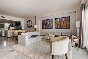 A modern living room with a sectional sofa, a round wall clock, abstract paintings, and a dining area with chairs. - Cosy Apartment inside Puerto Marina, steps to the Sea (Benalmádena)