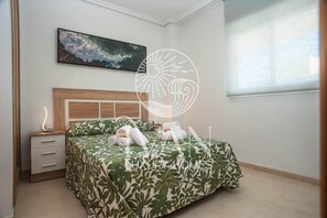 Hotellihuone, jossa on sänky, yöpöytä, seinälle asennettu televisio ja ikkuna, jossa on sälekaihtimet.