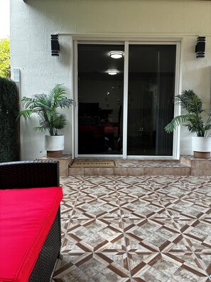 A covered patio area with a patterned tile floor, a red cushioned seating area, and potted plants. - LEIRA HOMES • Spacious 3BR Near Florida Keys • Free Parking (Florida City)