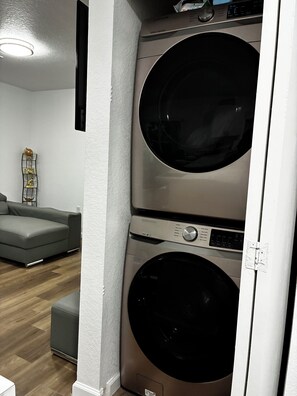 A laundry room with two stacked washing machines, a grey sofa, and a decorative shelf with figurines. - LEIRA HOMES • Spacious 3BR Near Florida Keys • Free Parking (Florida City)