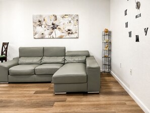 A modern living room with a grey sectional sofa, a decorative abstract painting, a metal sculpture, and a wall clock. - LEIRA HOMES • Spacious 3BR Near Florida Keys • Free Parking (Florida City)