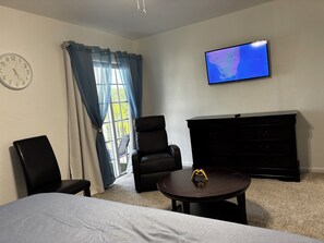 A hotel room with a bed, two chairs, a small table with a decorative item, a television on the wall, and a large window with curtains. - LEIRA HOMES • Spacious 3BR Near Florida Keys • Free Parking (Florida City)