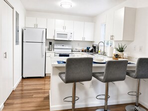 A modern kitchen with white cabinetry, a built-in microwave, a stainless steel refrigerator, a double sink, and a bar area with two stools. - LEIRA HOMES • Spacious 3BR Near Florida Keys • Free Parking (Florida City)