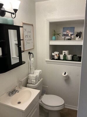 A bathroom with a white sink, toilet, and a mirror with a towel rack. There is a framed sign on the wall and a shelf with toiletries. - Fabulous 2-bedroom house with AC in charming River Oaks (River Oaks)