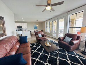 A spacious living room with a brown leather sofa, a coffee table, and a ceiling fan. A kitchen with white cabinets and a dining area with chairs are visible in the background. - Lake Travis Water Front - 180' Water Views  -3 beds and two baths (Jonestown)