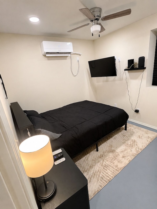 A bedroom with a bed, a desk with a lamp, a ceiling fan, an air conditioner, a mounted TV, and a wall-mounted shelf.