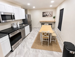 A compact kitchen with modern appliances, a dining area with a wooden table and chairs, and a small window with blinds.