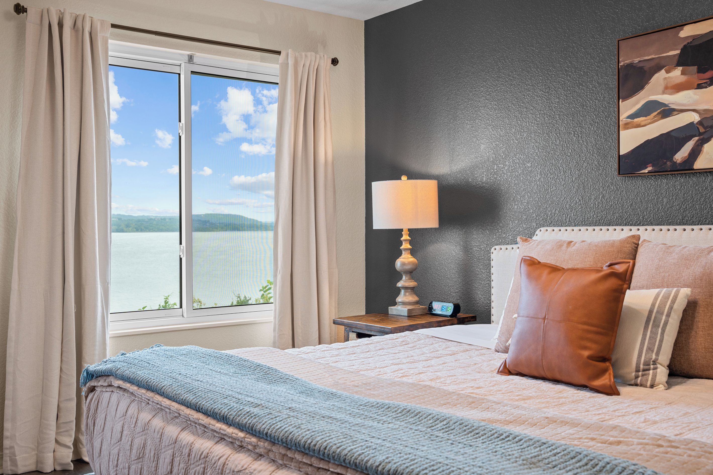 Ein Schlafzimmer mit großem Fenster, einem Bett mit blauer Decke, einer Nachttischlampe und einer Uhr auf einem Nachttisch.