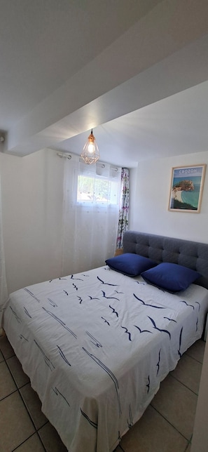 A bedroom with a bed, a window with curtains, a painting on the wall, and a hanging light fixture. - Vrbo Property (Saint-Évarzec)
