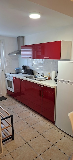 A kitchen with red cabinets, a white countertop, a stove, a sink, and a refrigerator. - Vrbo Property (Saint-Évarzec)