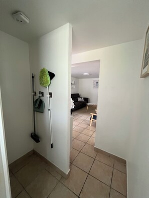 Ein Flur mit einem Mopp und einem Besen, die an der Wand lehnen, einem gefliesten Boden und einer Tür zu einem Wohnbereich mit einem Sofa und einem kleinen Tisch.