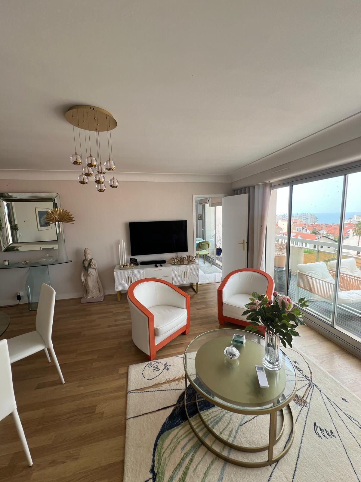 Un salon moderne doté d’un grand téléviseur, de deux fauteuils blancs ornés de bordures orange, d’une table basse en verre et offrant une vue sur la ville grâce à de grandes fenêtres.