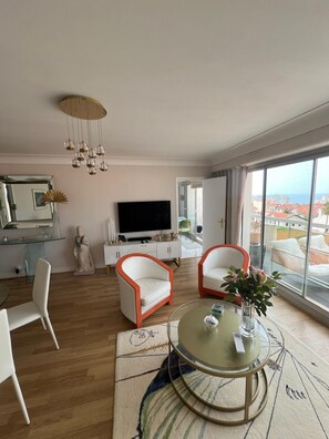 Uma sala de estar moderna com uma TV grande, duas cadeiras brancas com detalhes laranja, uma mesa de centro de vidro e vista da cidade através de grandes janelas.