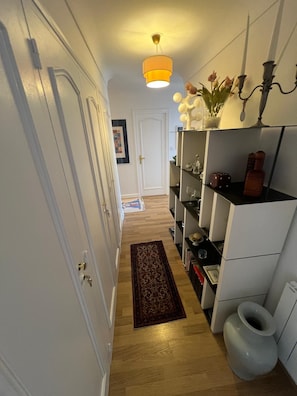 Um corredor com piso de madeira, tapete, porta branca e prateleira com diversos objetos.