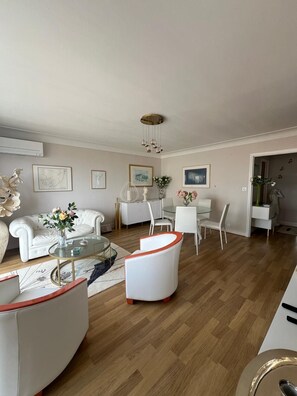 Uma sala de estar moderna com um sofá branco, uma mesa de centro de vidro e uma área de jantar com cadeiras brancas.