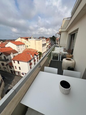 Ein Balkon mit einem weißen Tisch und einem weißen Topf, mit Blick auf eine Straße mit Gebäuden und roten Ziegeldächern.