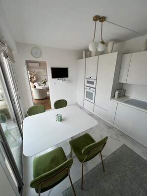 Una cocina moderna con armarios blancos, un televisor de pantalla plana y un comedor con mesa blanca y sillas verdes.