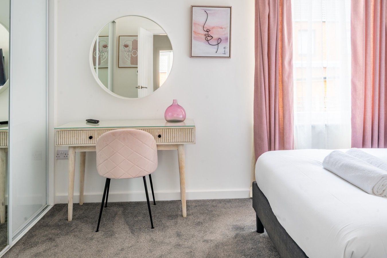 A bedroom with a bed, a desk with a chair, a round mirror, a pink vase, and a framed picture on the wall.