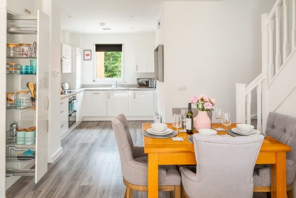 A modern kitchen with white cabinets, a dining area with a wooden table set for two, and a view of the outdoors through a window. - Broomsfield House |2 Bed | Parking |UEA | Hospital (Norwich)