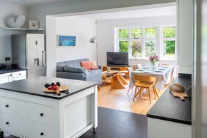 A modern kitchen with a central island, a dining area with a table set for four, and a living space with a sofa and television. - Swift Host | Scriveners | Blakeney | Sleeps 12 | Pet Friendly | Free Parking (Blakeney)