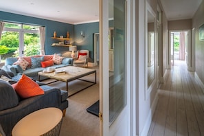 A modern living room with a sofa, coffee table, and a view of a garden through large windows. - Swift Host | Scriveners | Blakeney | Sleeps 12 | Pet Friendly | Free Parking (Blakeney)